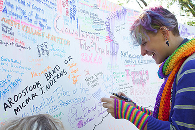 Junior art education student Sanja Petrashek writes 1 of 568 federally recognized tribe names on a mural next to the End of the Trail statue on campus to honor Indigenous People’s Day on campus Monday, Oct. 10. The structure is planned to stand until the end of the semester. (Photo by Allison Mueller)